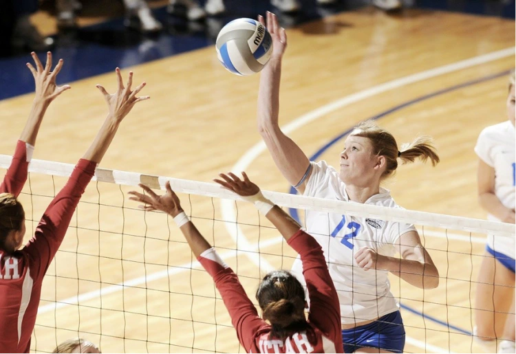 Frau im weißen Trikot beim Volleyballspiel, die den Ball über das Netz schlägt, während Spielerinnen des gegnerischen Teams versuchen zu blocken.